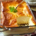 A glass baking dish holds a Ricotta Self-Filling Cake with a golden-brown crust. A slice is missing, revealing the creamy interior. A sprig of mint decorates the top, while a serving spatula rests beside it.