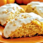 Three homemade pumpkin scones on an orange plate, boasting a golden-brown crumb and white icing atop. The background is softly blurred, beautifully highlighting the scones' texture and pumpkin-infused glaze.