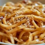 Close-up of homemade hibachi noodles drizzled with a glossy sauce, garnished with black and white sesame seeds and small bits of red chili peppers. The dish is served in a round, shallow bowl, capturing the essence of your favorite hibachi noodles recipe.