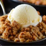 A bowl of warm apple crumble, reminiscent of an easy pumpkin pie crisp, is topped with a scoop of vanilla ice cream. The golden brown crumble pairs beautifully with the melting ice cream, adding a creamy contrast. A hint of cinnamon streusel elevates the flavors, with a spoon partially visible in the bowl.