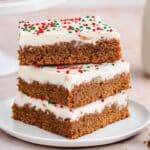 Three chewy gingerbread cookie bars stacked with white frosting and festive red and green sprinkles on a white plate, accompanied by a glass of milk. Perfect for your holiday cookie bar collection!