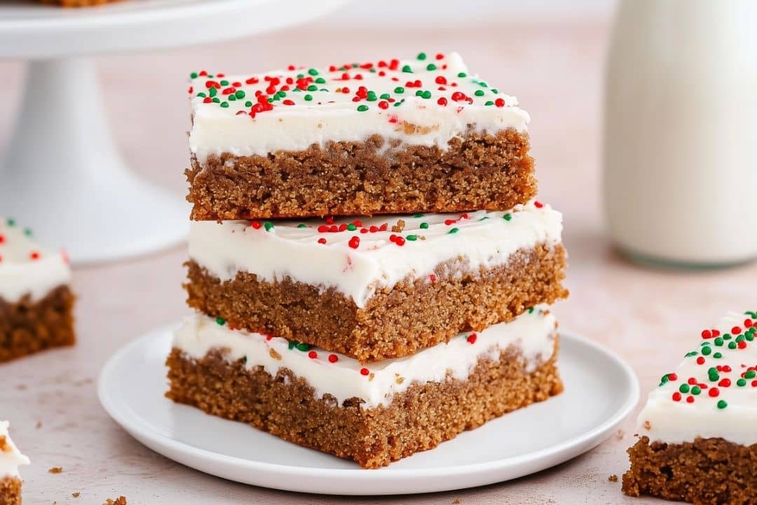 Three chewy gingerbread cookie bars stacked with white frosting and festive red and green sprinkles on a white plate, accompanied by a glass of milk. Perfect for your holiday cookie bar collection!