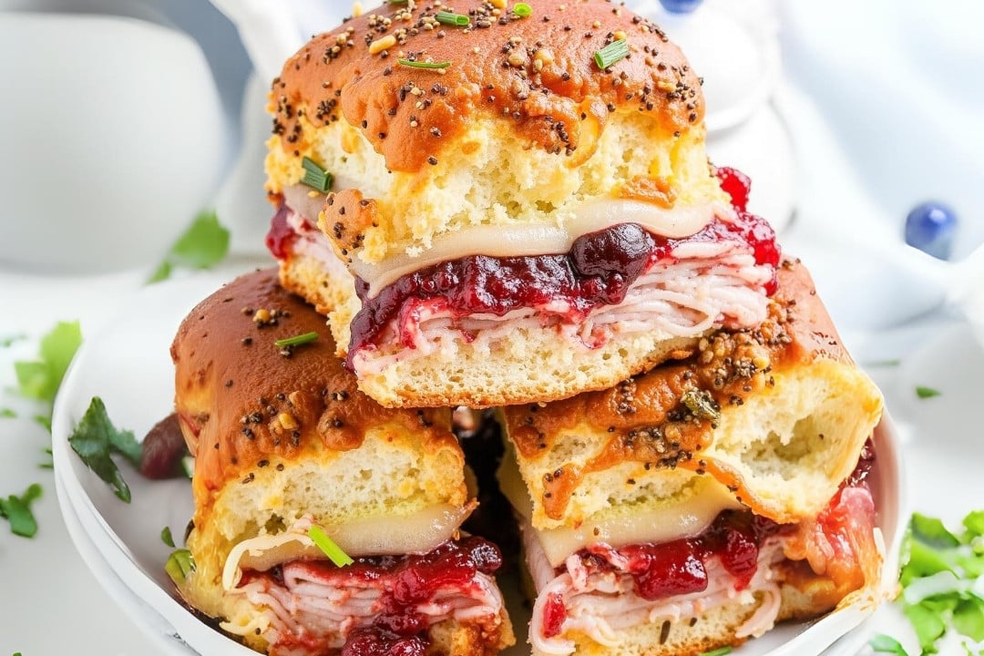 Three turkey and cranberry sliders stacked on a plate, featuring sliced turkey, melted cheese, and cranberry sauce nestled in poppy seed-topped Hawaiian rolls. This Cranberry Turkey Sliders recipe is garnished with chopped parsley and set against a white background with blueberries nearby.