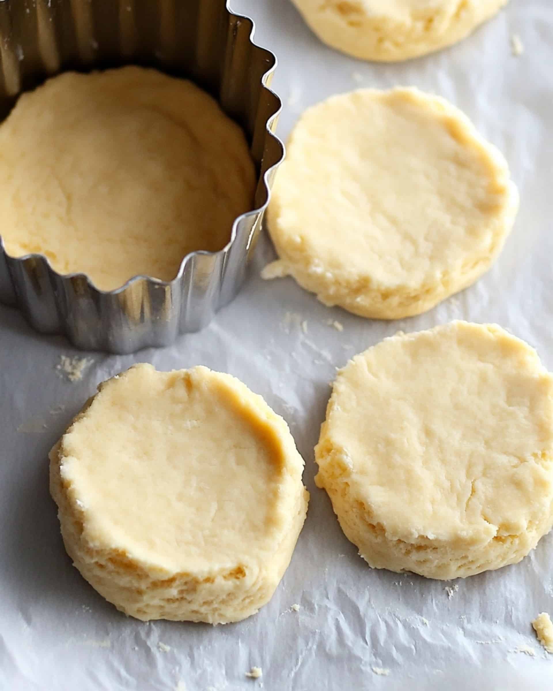 A top-down view of biscuit dough being cut on a floured marble surface. A metal biscuit cutter is pressing down into the pale yellow dough, creating a perfect circle. Three biscuit rounds have already been cut out, and three more are visible in the background, cooking in a black cast iron skillet. The dough appears soft and slightly textured with visible flour.