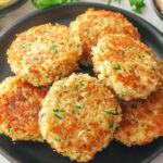 A black plate with six golden-brown crab cakes garnished with herbs. A lemon wedge, fresh parsley, and a small bowl of dipping sauce are visible in the background—perfect for those looking to try an air fryer crab cakes recipe.