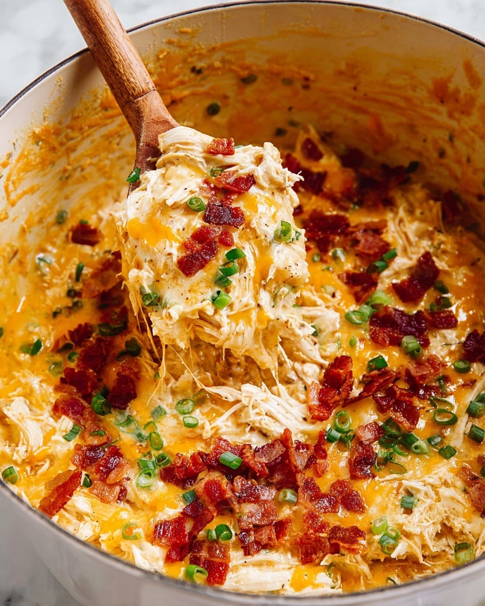 The image shows a creamy chicken bacon ranch dip in a white dish with a wooden spoon scooping some of the dip. The dip has shredded white chicken mixed in a thick, pale yellow sauce. The top layer is melted orange cheddar cheese with crispy reddish-brown bacon pieces scattered on top. Green slices of fresh green onion and small bits of parsley are sprinkled over the cheese and bacon, adding a touch of color. The inside edges of the dish have remnants of the creamy dip, giving a sense of texture. The scene is set on a white marbled surface. Photo taken with an iphone --ar 4:5 --v 7