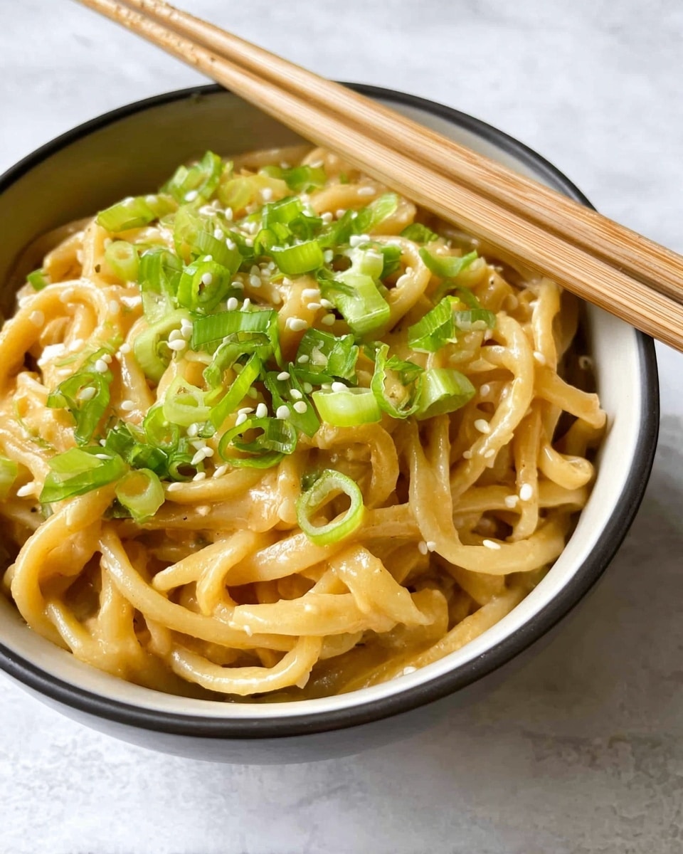 A close-up top view of a black bowl filled with thick, light brown noodles mixed with green sliced scallions and sesame seeds scattered on top. The noodles have a slight shine and soft texture, with small pieces of green vegetables throughout. The bowl sits on a white marbled surface, and a pair of light wooden chopsticks rests diagonally to the right of the bowl. photo taken with an iphone --ar 4:5 --v 7