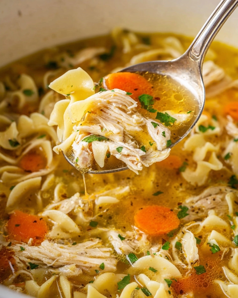 A close-up view of a spoon lifting a portion of chicken noodle soup from a bowl with a white marbled texture surface. The soup has multiple layers starting with a clear golden broth at the bottom, filled with shredded white chicken meat mixed with soft yellowish egg noodles. The soup also shows bright orange carrot slices mixed evenly among the chicken and noodles. Scattered small green parsley leaves add a fresh touch throughout the soup. The shiny silver spoon holds a mix of these ingredients with broth dripping off slightly, showing texture and color contrast. photo taken with an iphone --ar 4:5 --v 7