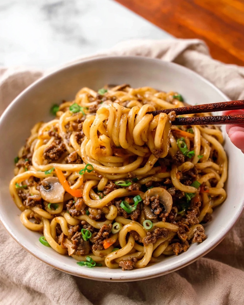 A bowl of thick, pale yellow noodles is shown with a woman's hand holding dark brown chopsticks lifting some noodles above the bowl. The noodles are mixed with small pieces of cooked ground meat that are dark brown and bits of green onion and thin orange carrot strips scattered throughout. The bowl is white and sits on a white marbled surface, with green leaves blurred in the background. The noodles look soft and slightly shiny with a light sauce. photo taken with an iphone --ar 4:5 --v 7