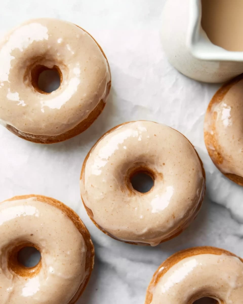 Pumpkin Donuts with Maple Glaze Recipe 5 The image shows a group of round donuts with a smooth, light beige glaze on top that has a slightly shiny texture. Each donut has a visible hole in the center. The donuts are placed on a white marbled surface, with some of their edges softly touching each other. In the top right corner, part of a white bowl or cup with a liquid inside is visible. The overall scene has natural lighting that highlights the soft texture of the glaze and the light golden-brown color of the donuts underneath. photo taken with an iphone --ar 4:5 --v 7
