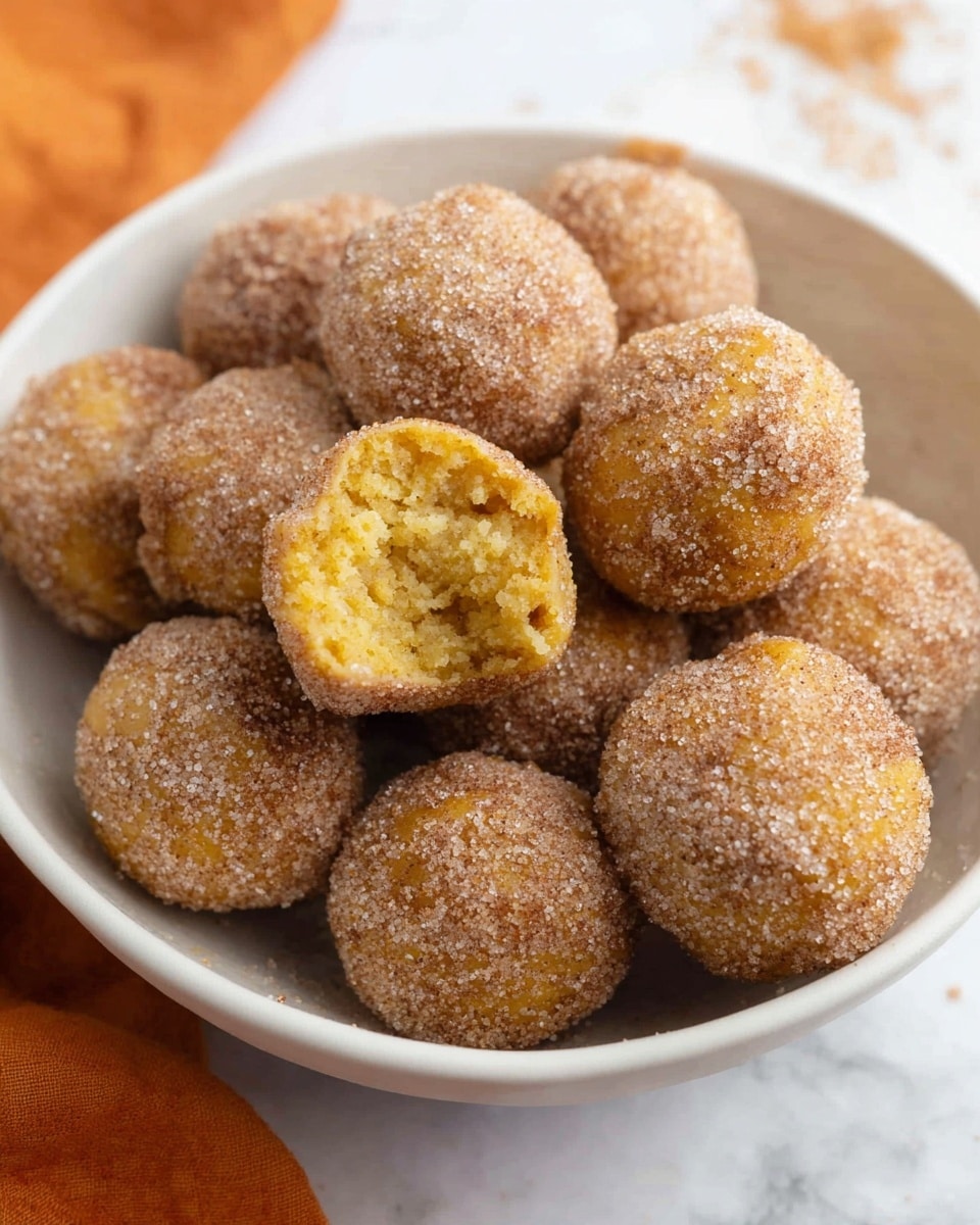 Easy Bisquick Pumpkin Donut Holes Recipe 6 A white oval plate with a thin gold rim holds twenty small round balls arranged closely together in a loose triangular shape. Each ball is golden-brown and coated evenly with a grainy layer of cinnamon sugar, giving a slightly rough texture. Around the plate, three cinnamon sticks and a larger cinnamon piece lie on the white marbled surface. The top left corner shows a blurred bunch of orange reeds in a white vase, with a soft orange and white striped cloth partially visible at the bottom left of the image. The lighting is soft and warm, highlighting the texture and color of the balls. photo taken with an iphone --ar 4:5 --v 7