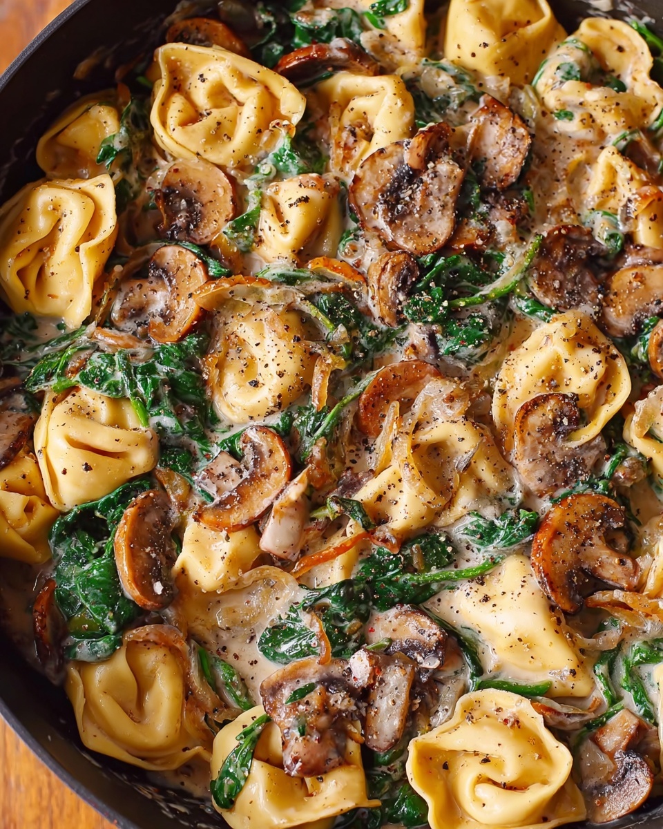 The image shows a close-up of a pan filled with soft yellow tortellini pasta mixed with sautéed mushrooms that have a brown and creamy texture, wilted dark green spinach leaves, and bits of light brown caramelized onions. The dish is covered in a creamy white sauce with specks of black pepper sprinkled all over, giving the meal a seasoned look. The pan sits on a wooden surface, but the main focus is the creamy, colorful, and textured mix inside the pan. photo taken with an iphone --ar 4:5 --v 7