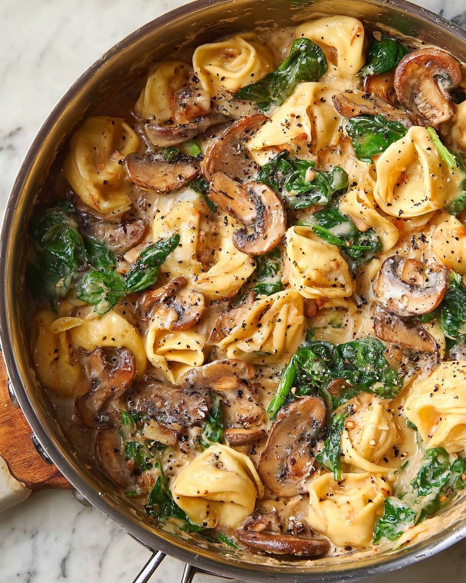 A close-up view of a large stainless steel pan filled with creamy sauce layered with golden yellow tortellini, sliced brown mushrooms, and bright green spinach leaves, all mixed together and sprinkled with coarse black pepper. The creamy sauce has a light beige color, smooth texture, and pools around the pasta and vegetables. The tortellini appear soft and slightly shiny, while the mushrooms have a tender, cooked look with some browned edges. The spinach adds a fresh, leafy green contrast, and the black pepper specks are scattered evenly over the whole dish. The background is a white marbled surface. photo taken with an iphone --ar 4:5 --v 7