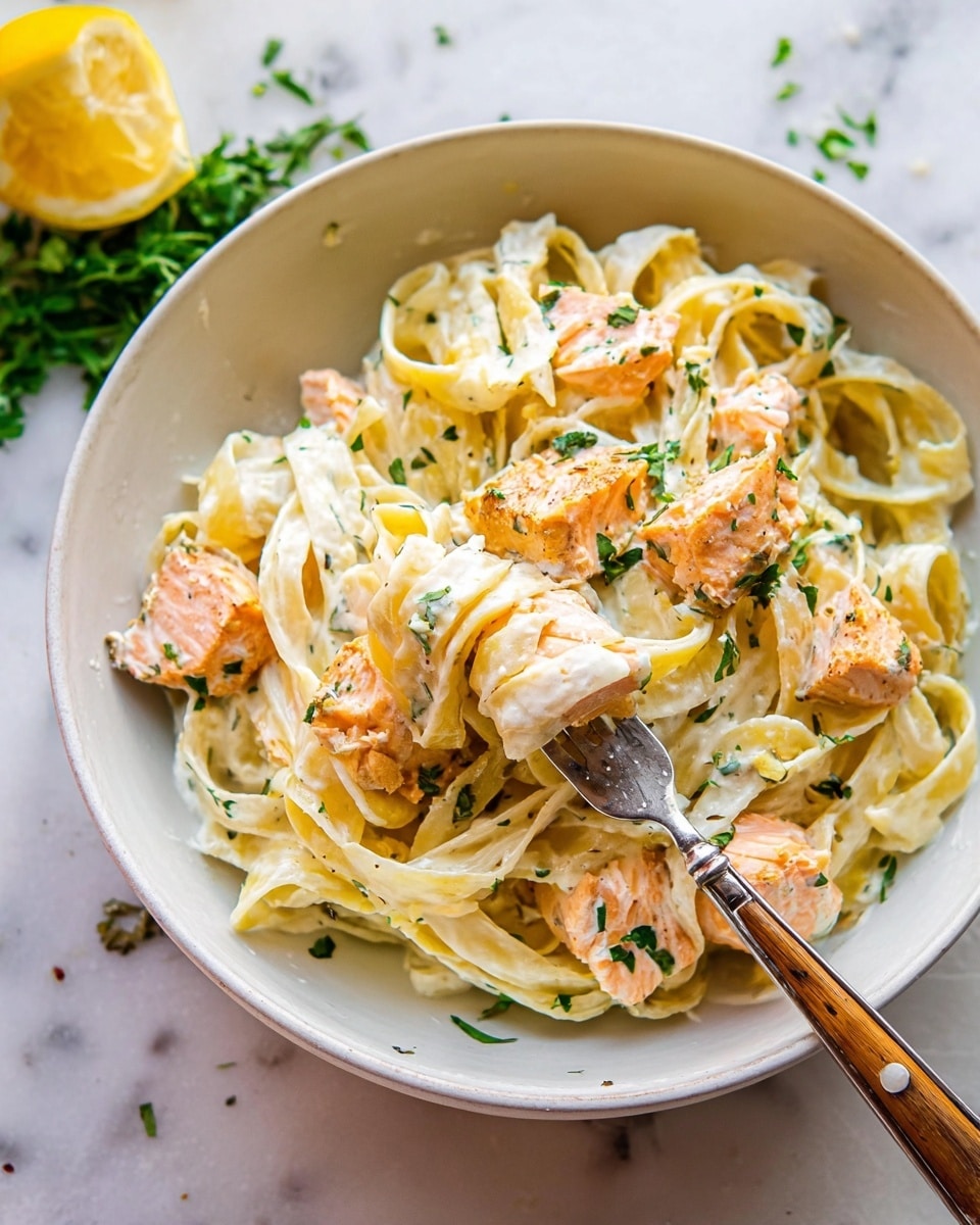A white bowl holds a creamy pasta dish with three layers visible: at the bottom is a smooth white cream sauce, on top of that are long, flat pale yellow noodles spread throughout, and scattered across the noodles are pinkish-orange chunks of cooked salmon with light brown seared edges. Mixed in among the pasta and salmon are bright green leafy pieces, adding freshness and contrast to the dish. The creamy sauce clings to the noodles and salmon, making the whole look rich and smooth. Photo taken with an iphone --ar 4:5 --v 7