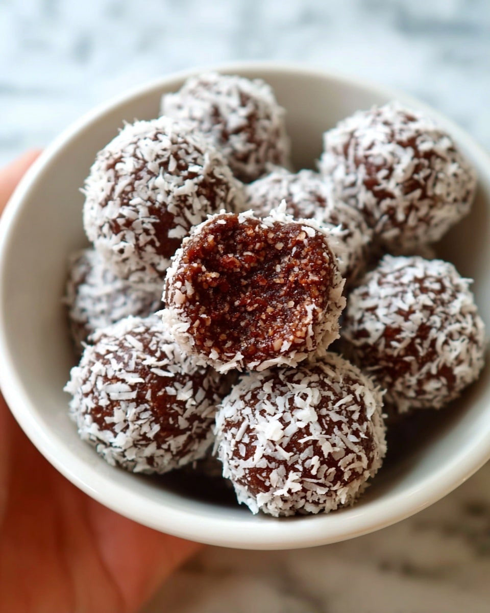 A woman's hand is holding a dark brown round ball with a rough texture, coated with white shredded coconut flakes. The ball is above a white plate filled with a thick layer of white shredded coconut, spread in a circular shape. More dark brown balls with a similar texture are scattered on the upper part of the plate. The plate is placed on a white marbled surface. photo taken with an iphone --ar 4:5 --v 7