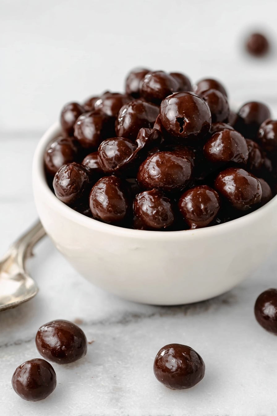 Chocolate Espresso Bean Bites Recipe 6 A small white bowl filled with shiny, dark brown chocolate-coated nuts piled high inside. The nuts have a smooth and glossy texture with some cracks showing their natural shape. Around the bowl on a white marbled surface, a few more chocolate-coated nuts are scattered, with some resting on a silver spoon at the bottom left corner. Photo taken with an iphone --ar 2:3 --v 7 - Chocolate Espresso Bean Bites, Chocolate Espresso Candy, Espresso Bean Snacks, Homemade Chocolate Espresso Treats, Easy Espresso Bites