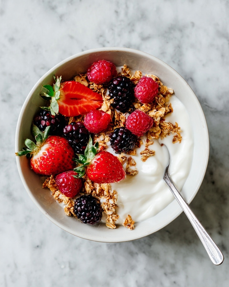 A white bowl filled with three main layers: at the bottom is creamy white yogurt, swirled on top with deep purple berry sauce, giving a marbled look. The top layer has crunchy golden granola covering about half the bowl, and fresh red strawberries, some whole and some sliced in half showing their bright red inside with green leaves still attached, scattered on the yogurt and sauce side. A dark spoon is placed inside the bowl near the strawberries. The bowl sits on a white marbled surface. photo taken with an iphone --ar 4:5 --v 7