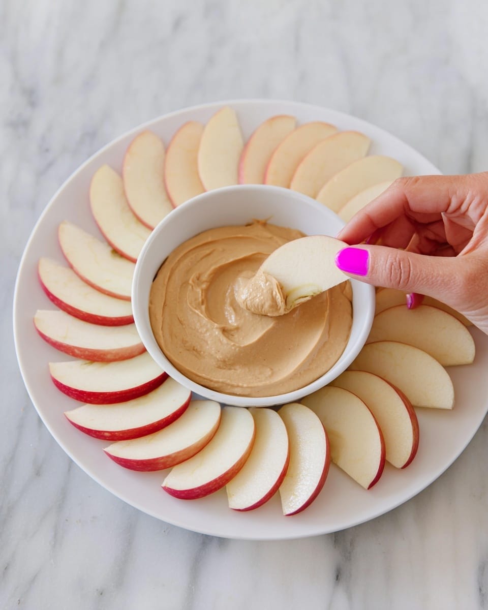 A white plate holds a white bowl filled with a smooth, light brown creamy dip. Around the bowl, there is a single layer of apple slices, each slice pale cream with light red edges, arranged in a neat circle. A woman's hand with bright pink nail polish is lifting an apple slice, ready to dip it into the creamy spread. The plate sits on a white marbled texture surface. photo taken with an iphone --ar 4:5 --v 7