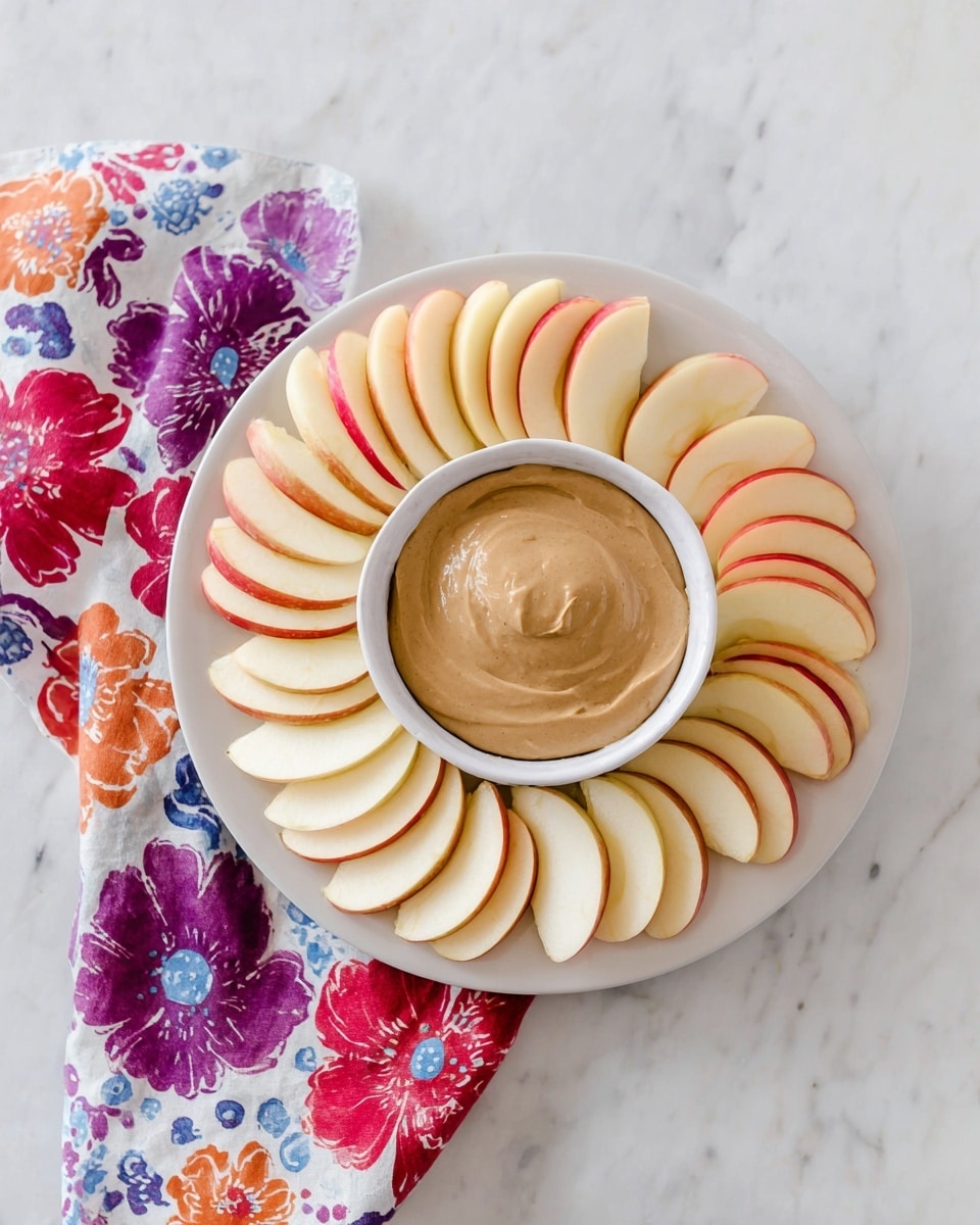 A white plate is filled with two layers of apple slices arranged in a circular pattern. The apple slices on the outside have a pale cream color with red edges, and the inner layer of slices shows darker red skin with a pale side. In the center of the plate, a white bowl holds a thick, smooth, light brown dip with soft swirls on the surface. The plate is set on a white marbled surface, and a colorful cloth with blue, black, and red spots is partially visible on the left side. photo taken with an iphone --ar 4:5 --v 7