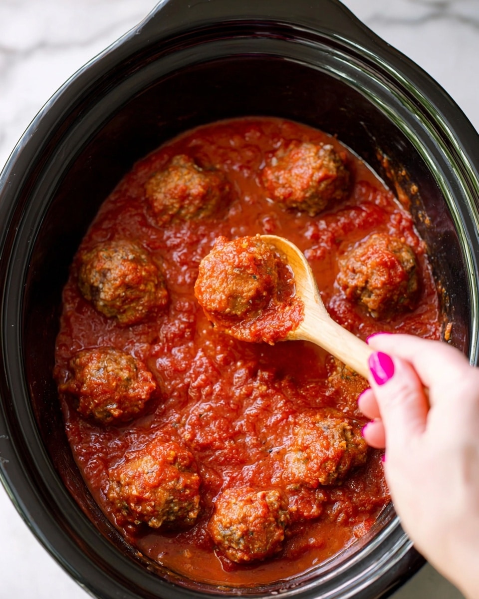 The image shows a black slow cooker filled with a thick, textured red tomato sauce covering nine round, brown meatballs. A woman's hand with pink painted nails is holding a wooden spoon that lifts one meatball, showing its moist, uneven surface coated in sauce. The red sauce has bits of herbs and a slightly chunky texture, spreading evenly inside the slow cooker. The background is a white marbled surface. photo taken with an iphone --ar 4:5 --v 7