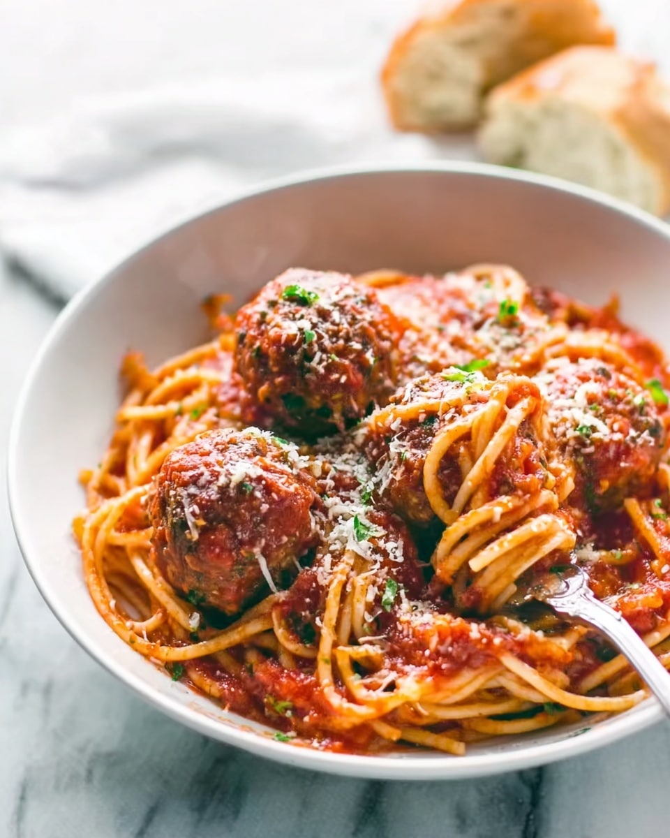A white bowl filled with spaghetti covered in red tomato sauce, topped with three large brown meatballs sprinkled with grated white cheese and small green herb bits. A silver fork rests inside the bowl on the right side, curling some noodles. The background shows a white marbled surface with some bread in soft focus. The image is bright and clear, capturing the textures of the sauce, meatballs, and cheese. Photo taken with an iphone --ar 4:5 --v 7