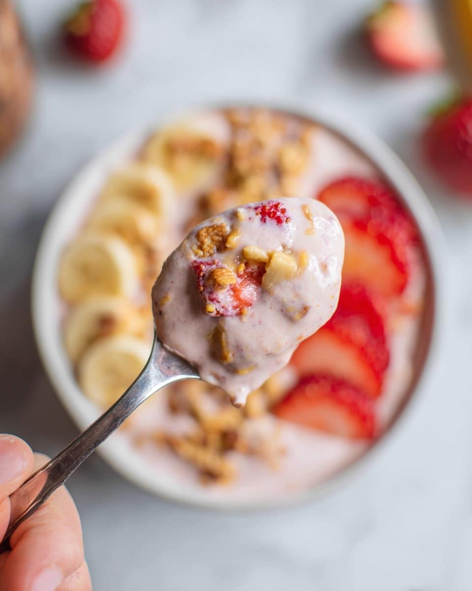 Strawberry Banana Smoothie Bowl Recipe 6 A close-up image of a metal spoon held by a woman's hand, filled with a light pink creamy mixture containing small pieces of red strawberries, tan nut chunks, and small bits of granola. In the blurred background, a white bowl with a similar pink base mixture can be seen, layered with bright red strawberry slices on one side, light beige banana slices on another, and sprinkled with golden brown granola on top. The whole scene is set on a white marbled surface. photo taken with an iphone --ar 4:5 --v 7