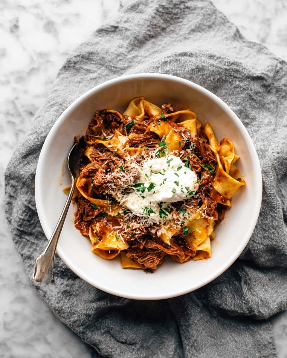 Slow Cooker Beef Ragu with Pappardelle Recipe 5 A white bowl holds a serving of wide, flat pasta ribbons in a rich brown-red sauce with shredded meat mixed throughout. On top, there is a dollop of creamy white cheese or cream, sprinkled with grated cheese and chopped green herbs. A silver spoon rests in the bowl on the left side. The bowl is placed on a wrinkled gray cloth over a white marbled surface. photo taken with an iphone --ar 4:5 --v 7
