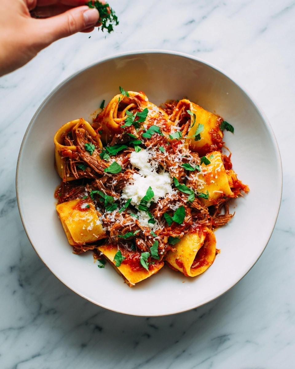 Slow Cooker Beef Ragu with Pappardelle Recipe 6 A white bowl on a white marbled surface holds a generous serving of rolled pasta mixed with shredded meat and covered in red sauce. On top of the pasta, there are fresh green herb leaves scattered, adding a bright color contrast. A small dollop of white cheese rests on one side of the pasta. A woman's hand is visible at the top of the image, sprinkling more herbs onto the dish. Photo taken with an iphone --ar 4:5 --v 7