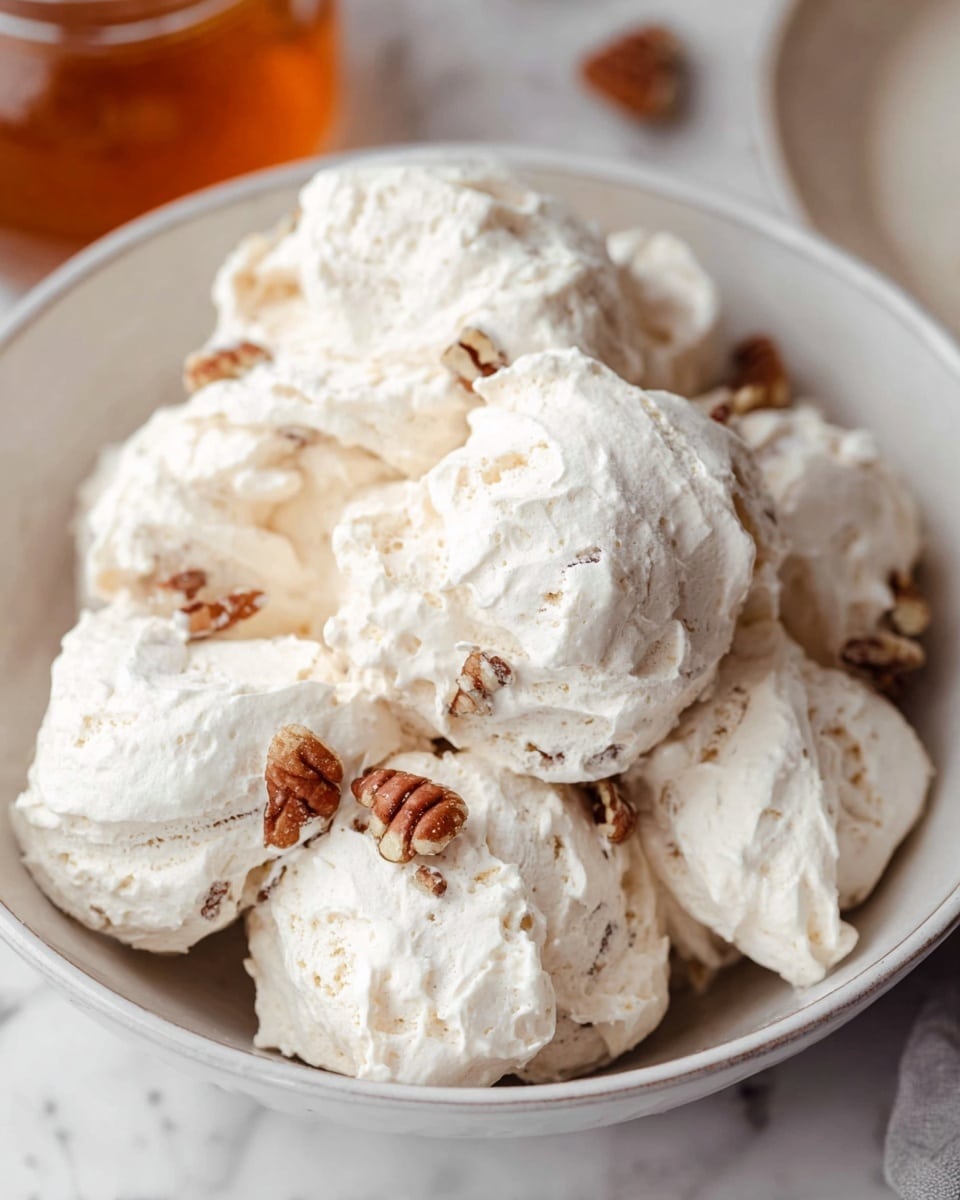 A white bowl is filled with about ten irregularly shaped scoops of light cream meringue, each scoop textured with soft peaks and small rough edges. Mixed inside the meringue are small pieces of pecan nuts, showing a few whole pecans around the scoops, adding brown contrast to the pale cream. The bowl sits on a surface with a white marbled texture. The image focuses closely on the bowl, with a slight view of another dish and a jar of amber liquid partially blurred in the background, photo taken with an iphone --ar 4:5 --v 7