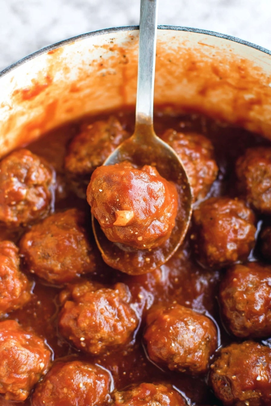 This image shows many small, round meatballs covered in a thick reddish-brown sauce. The meatballs have a slightly shiny and moist texture, with some small bits of seasoning visible on their surface. They sit closely together in a pot with sauce splattered along the inside edges. A silver spoon lifts two meatballs above the others, and the sauce clings to both the meatballs and the spoon. The background has a white marbled texture. photo taken with an iphone --ar 2:3 --v 7 - Cranberry BBQ Meatballs, easy meatball appetizer, slow cooker meatball recipe, sweet and tangy meatballs, holiday party snacks