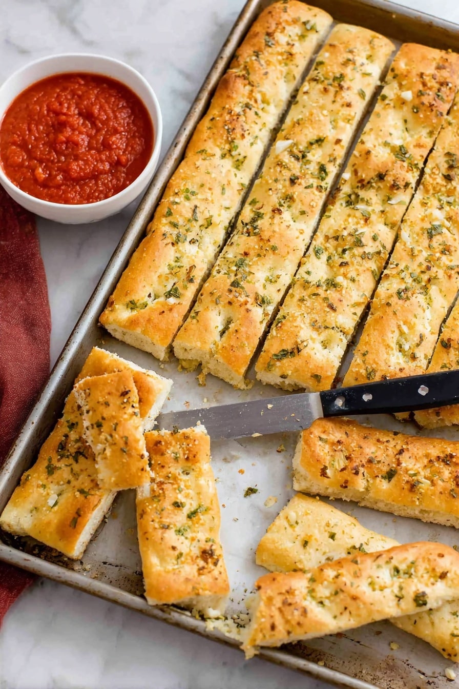 Copycat Pizza Hut Breadsticks Recipe 8 The image shows a metal baking tray filled with rectangular, golden-brown breadsticks, each sprinkled with green herbs and bits of garlic on top. The breadsticks are arranged in three rows inside the tray, and a few pieces are placed outside the tray on a white marbled surface. A knife with a black handle rests on the breadsticks inside the tray. Next to the tray, there is a white bowl filled with red marinara sauce, with one breadstick partially dipped in it. The breadsticks look soft inside and slightly crispy on the outside. Photo taken with an iphone --ar 2:3 --v 7 - Copycat Pizza Hut Breadsticks, homemade Pizza Hut breadsticks, how to make Pizza Hut breadsticks, best breadsticks recipes, cheesy garlic breadsticks