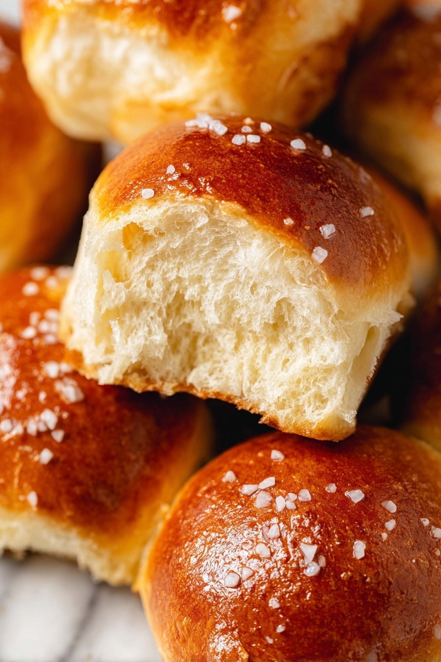 The image shows a close-up view of soft, golden brown bread rolls with a shiny crust topped with coarse salt crystals. One roll is torn open in the center, revealing a fluffy, light inside with a soft texture. The rolls have a slightly uneven surface with a warm, inviting color. They rest against each other on a white marbled surface, creating a cozy and appetizing arrangement. photo taken with an iphone --ar 2:3 --v 7 - Homemade Pretzel Rolls, pretzel rolls recipe, soft pretzel rolls, warm pretzel buns, easy pretzel bread