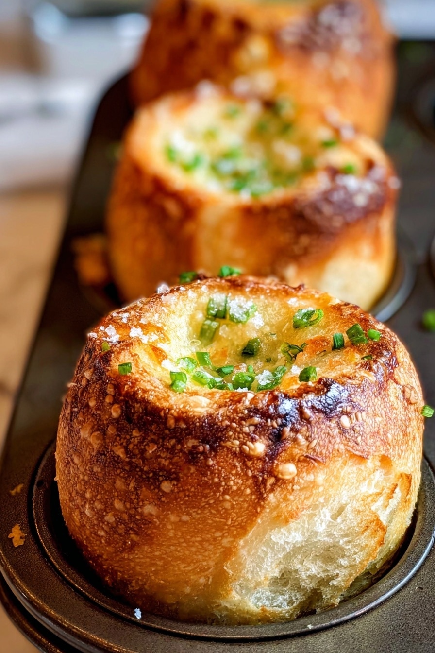 Pancetta Parmesan Popovers Recipe 6 The image shows three golden brown baked rolls in a close-up view, each roll standing upright with a hollow center filled with a light grilled texture topped with small green chive pieces and coarse white salt. The rolls have a thick, crispy outer crust with a soft, airy inside visible through small cracks. They are placed in a dark metal tray with individual compartments for each roll. The background and surface are a soft, blurred white marbled texture. photo taken with an iphone --ar 2:3 --v 7 - Pancetta Parmesan Popovers, savory popovers with pancetta and Parmesan, cheesy crispy popover recipe, easy savory breakfast bites, baked pancetta appetizer