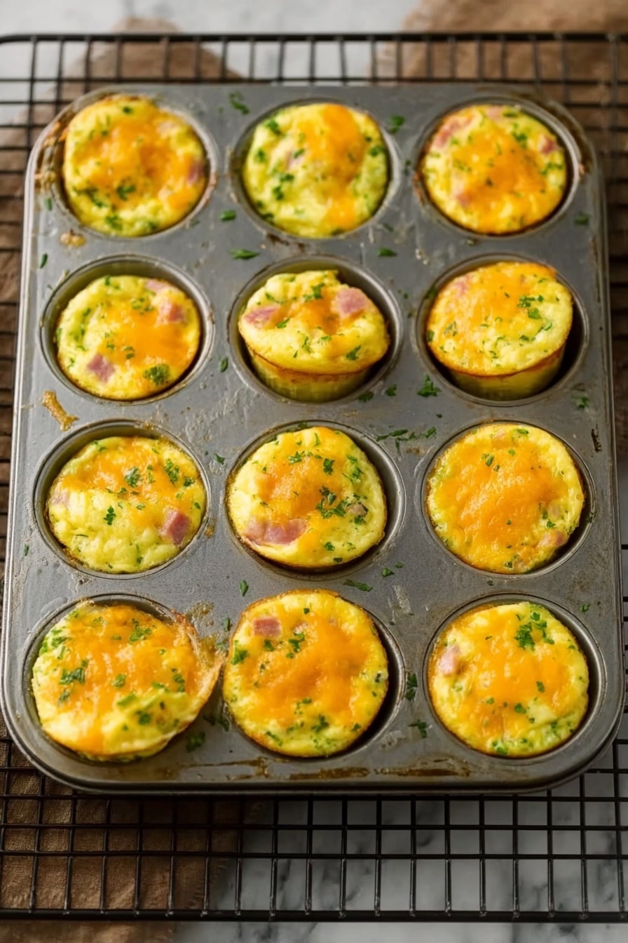 A metal muffin pan holds 12 small egg muffin cups, each filled with layers of fluffy yellow egg mixed with bits of green vegetables and pink ham. The muffins have a light golden top with melted orange cheese, creating a slightly bumpy and soft texture. Some muffins puff up more than others, showing a thick and airy inside with green herbs spread throughout. The pan rests on a rectangular black wire rack on a white marbled surface. Photo taken with an iphone --ar 2:3 --v 7 - Ham and Cheese Egg Muffins, Breakfast Egg Muffins, Quick Breakfast Recipes, Protein-Packed Breakfasts, Easy Egg Muffin Cups