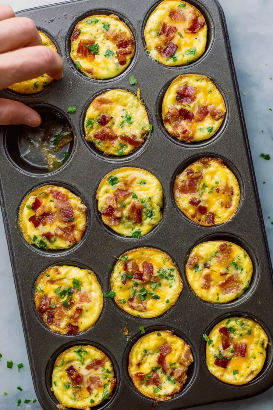 Cheddar Bacon Egg Muffins Recipe 6 The image shows a dark metal muffin tray filled with twelve small egg bites. Each bite has a bright yellow egg base mixed with pieces of cooked bacon that have a reddish-brown color and small bits of green herbs or vegetables scattered on top and inside. The egg bites are slightly raised and have a soft, slightly shiny texture with some cheese melted and golden brown on top. The tray rests on a white marble surface, with a woman’s hand reaching toward the top left egg bite. Some small green herb pieces are scattered around the tray. photo taken with an iphone --ar 2:3 --v 7 - Cheddar Bacon Egg Muffins, breakfast egg muffins, bacon and cheese egg cups, grab-and-go breakfast recipes, easy egg muffin recipes