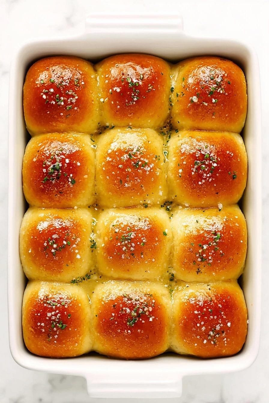A white rectangular baking dish holds twelve golden brown dinner rolls arranged in a 3 by 4 grid. Each roll is smooth, shiny, and slightly rounded on top, with a crust that is a warm golden color, showing a gentle gradient from darker brown centers to lighter edges. The tops of the rolls are sprinkled with coarse salt and small green herb flakes, adding texture and color contrast. The rolls sit closely together, with the sides touching, inside the clean white dish. The surface underneath is a white marbled texture. photo taken with an iphone --ar 2:3 --v 7 - Rosemary Garlic Dinner Rolls, homemade dinner rolls, quick bread recipes, savory bread recipes, fluffy dinner rolls