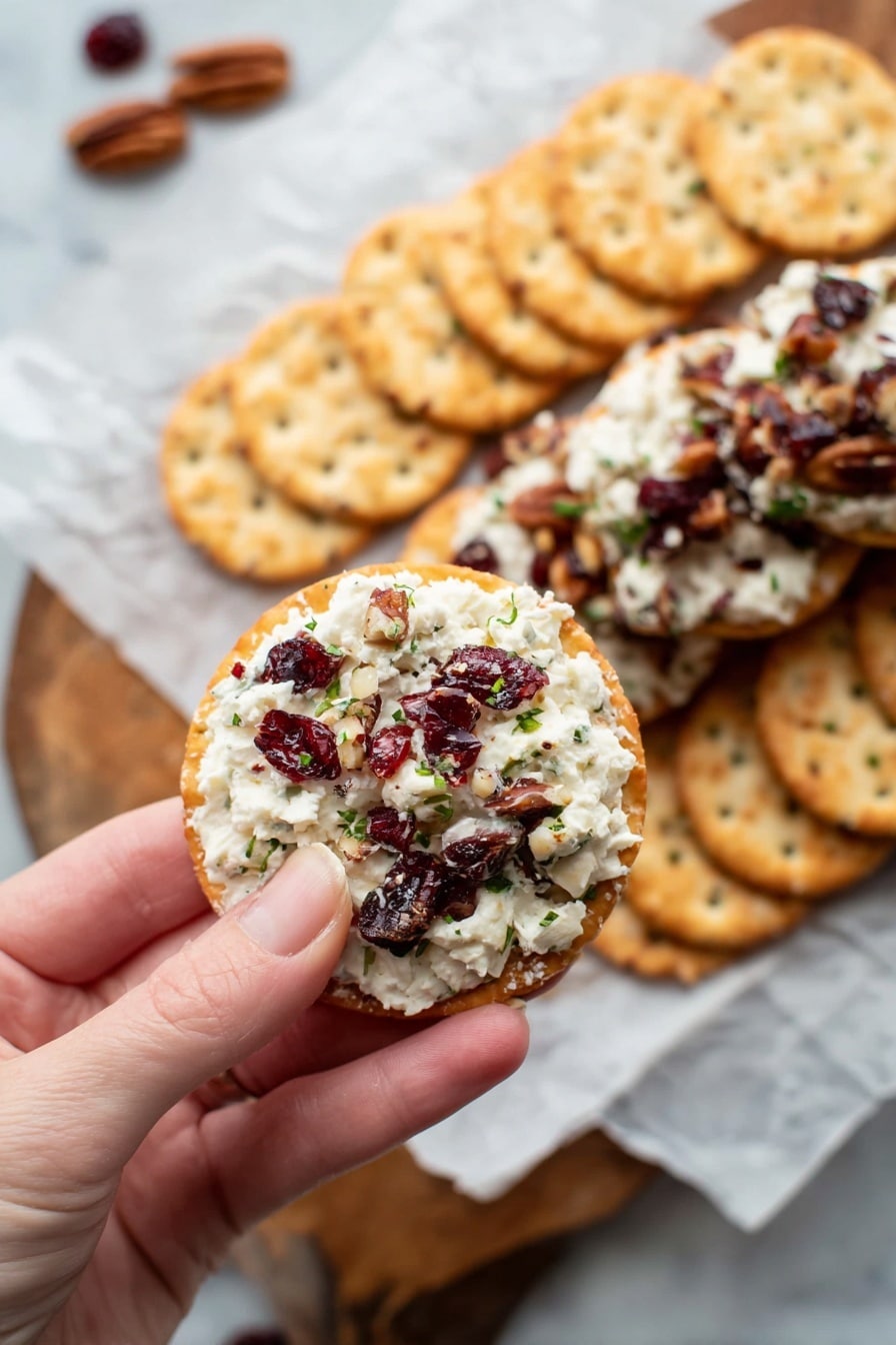 A log-shaped cheese ball covered with chopped nuts and pieces of dark red dried cranberries is shown close-up, resting on white parchment paper. The cheese inside is creamy white, mixed with bits of green herbs and pieces of cranberries and nuts. The outer surface of the log is rough with visible pieces of nuts and cranberries in warm brown, dark red, and green colors. Some crackers and scattered nut pieces appear blurred in the foreground on a white marbled surface. Photo taken with an iphone --ar 2:3 --v 7 - Cranberry Pecan Cheese Log, Cranberry Pecan Cheese Log, festive cheese appetizer, easy cheese log recipe, holiday party appetizer