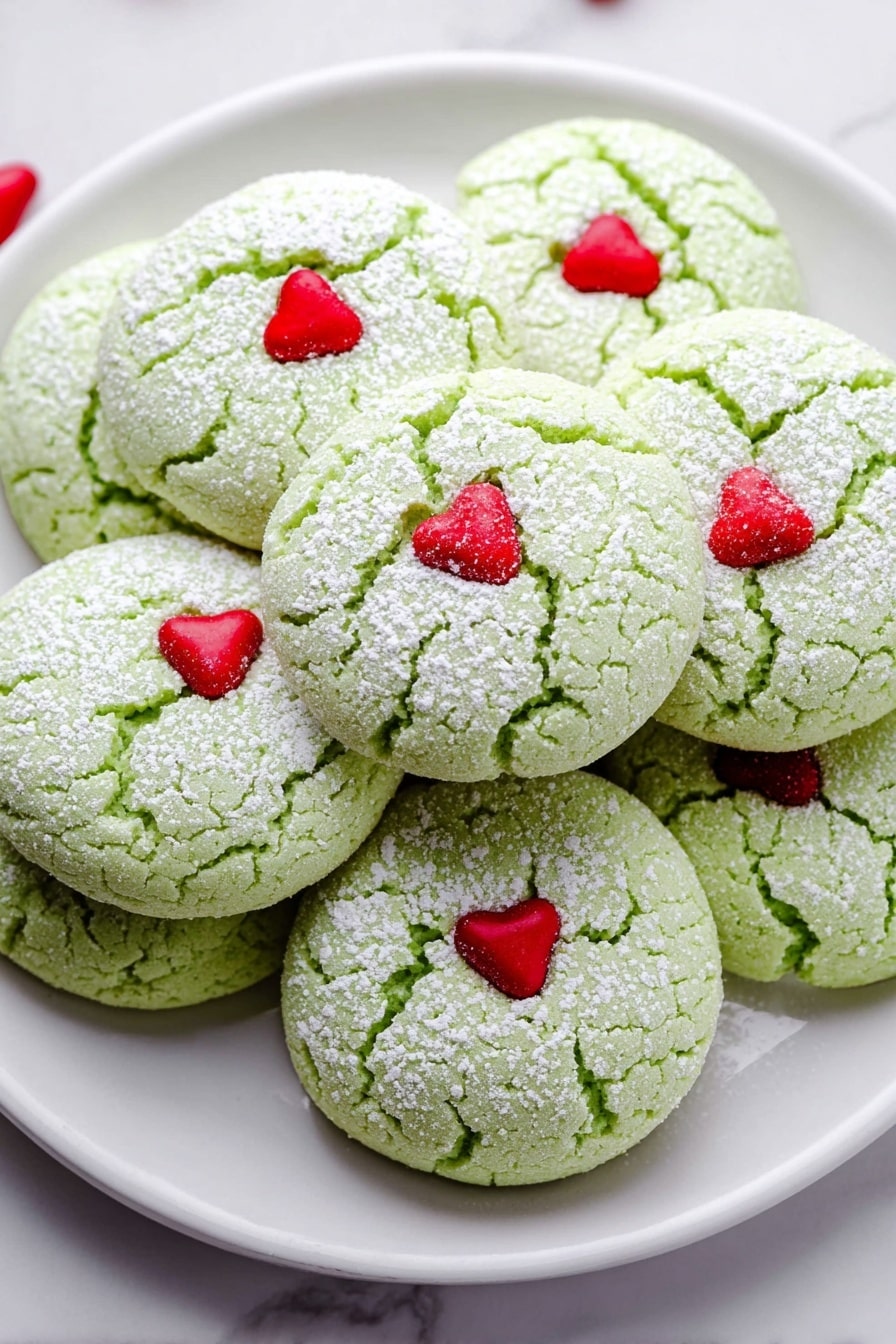 A white plate filled with multiple round, pale green cookies that have a cracked surface dusted with white powdered sugar. Each cookie is decorated with a small, red, heart-shaped candy placed near the center. The cookies are stacked close together on a white marbled surface, creating a soft, textured look. photo taken with an iphone --ar 2:3 --v 7 - Grinch Peppermint Cookies, peppermint holiday cookies, green Christmas cookies, easy festive cookie recipe, minty holiday treats