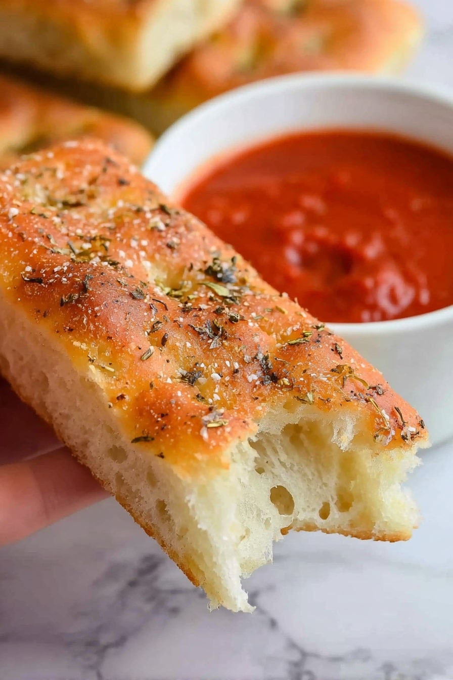 Copycat Pizza Hut Breadsticks Recipe 6 A close-up of a woman's hand holding a rectangular piece of focaccia bread with a light golden crust on top sprinkled with coarse salt and dried herbs, showing the soft, airy white inside at the bitten end. In the blurry background, a white bowl filled with thick red marinara sauce is visible, alongside another piece of focaccia bread partly visible on a white marbled surface. photo taken with an iphone --ar 2:3 --v 7 - Copycat Pizza Hut Breadsticks, homemade Pizza Hut breadsticks, how to make Pizza Hut breadsticks, best breadsticks recipes, cheesy garlic breadsticks