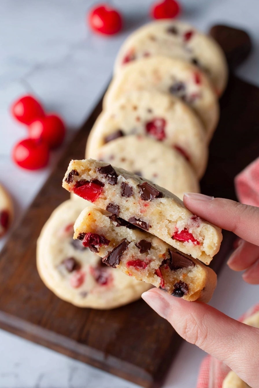A woman's hand holds a cookie broken in half, showing a soft, pale beige inside with bright red cherry pieces and dark brown chocolate chunks spread throughout. Behind, a row of six whole round cookies with similar colors and textures is arranged on a dark wooden board on top of a white marbled surface. Bright red cherries are placed near the cookies in the background. The cookie surface looks smooth with bits of cherry and chocolate visible. Photo taken with an iphone --ar 2:3 --v 7 - Maraschino Cherry Shortbread Cookies, holiday shortbread cookies, cherry buttery cookies, easy festive cookies, cherry shortbread recipe