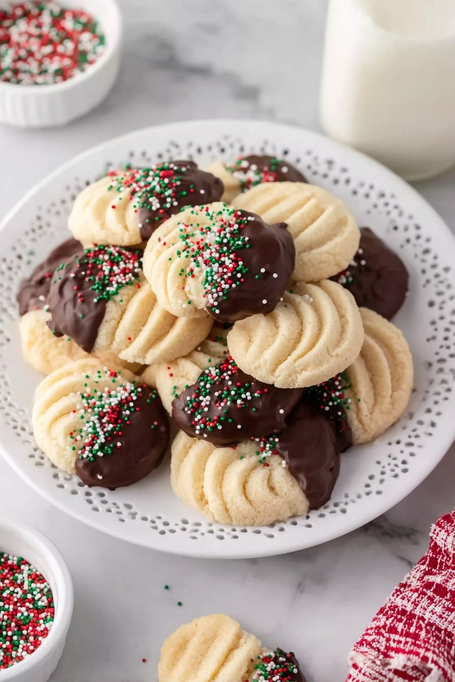Buttery Swirl Cookies with Chocolate Dip Recipe 8 A white ornate plate holds a pile of round, light beige cookies with a swirled texture, each partially dipped in dark chocolate on one side. The chocolate-dipped portions are decorated with small red, green, and white sprinkles. Around the plate, a few extra cookies lie on a white marbled surface, with a small white bowl of mixed color sprinkles and a glass bottle of milk nearby. A woman's hand can be seen holding one cookie on the right side. photo taken with an iphone --ar 2:3 --v 7 - Buttery Swirl Cookies with Chocolate Dip, elegant swirl cookies, chocolate dipped butter cookies, holiday cookie ideas, easy festive cookie recipes