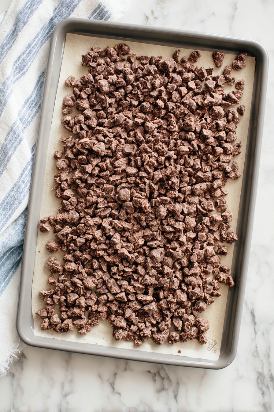 A gray baking tray lined with light beige parchment paper is filled with small, rough clusters of chocolate-colored pieces spread all over. The clusters are uneven in size, with some bigger chunks mainly on the right side and smaller clusters covering the rest. The tray sits on a white marbled surface with a soft blue and white striped cloth peeking out from the left side. The overall look is rustic and textured, showing a mix of smooth and bumpy surfaces on the chocolate clusters. photo taken with an iphone --ar 2:3 --v 7 - Reindeer Poop Snack Mix, holiday snack mix, festive Christmas treats, easy holiday recipes, chocolate holiday snacks