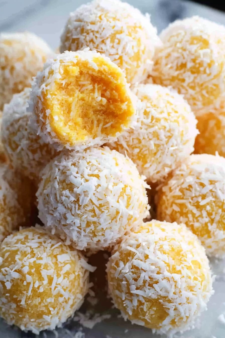 The image shows many round yellow-orange balls covered in white shredded coconut. One ball in the front is bitten, revealing a soft, moist, and textured inside that looks like a mix of grated ingredients. The balls are piled together on a white marbled surface, giving a sense of freshness and softness. The shredded coconut adds a fluffy, fine texture on the outside of each ball. photo taken with an iphone --ar 2:3 --v 7 - Pineapple Coconut Christmas Balls, tropical holiday treats, no-bake Christmas desserts, festive pineapple coconut bites, holiday candy balls