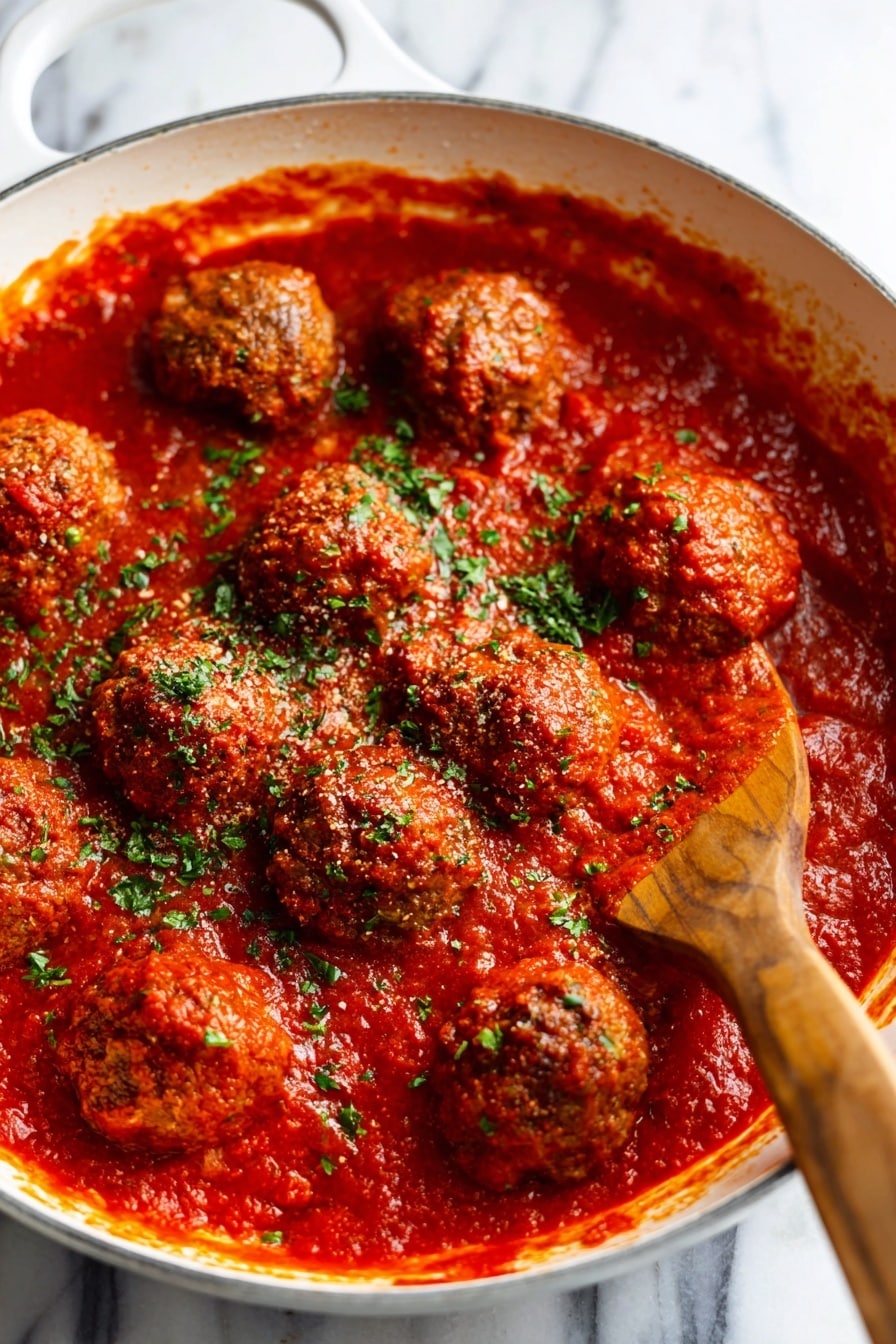 Cranberry Meatballs Recipe 8 The image shows a close-up of meatballs covered in thick red tomato sauce inside a white pan. The sauce is rich and smooth with a glossy texture, and the meatballs are round with a slightly crispy surface peeking through the sauce layer. Bright green chopped herbs are sprinkled evenly over the top, adding a fresh pop of color. A wooden spatula is partially visible on the right side, touching the sauce and meatballs. The background is a white marbled texture. photo taken with an iphone --ar 2:3 --v 7 - Cranberry Meatballs, holiday meatballs with cranberry sauce, easy savory meatball recipes, quick appetizer ideas, flavorful pork meatballs