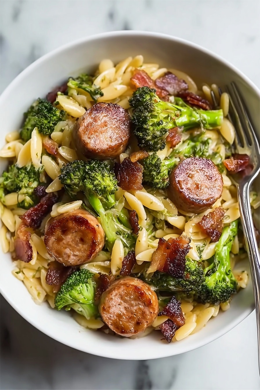The dish is served in a white bowl on a white marbled surface. It has a base layer of small, oval-shaped pasta in a light yellow color. Scattered throughout and on top are green broccoli florets with a slightly rough texture. On top of these are several round slices of browned sausage with a crispy, golden-brown outer surface and a juicy interior. Small pieces of cooked bacon or similar meat are mixed in, adding dark reddish-brown spots throughout the dish. A fork is placed inside the bowl on the right side. Photo taken with an iphone --ar 2:3 --v 7 - Creamy Chicken Sausage Broccoli Orzo, chicken sausage orzo dinner, easy creamy orzo recipes, hearty chicken sausage pasta, quick weeknight meal ideas