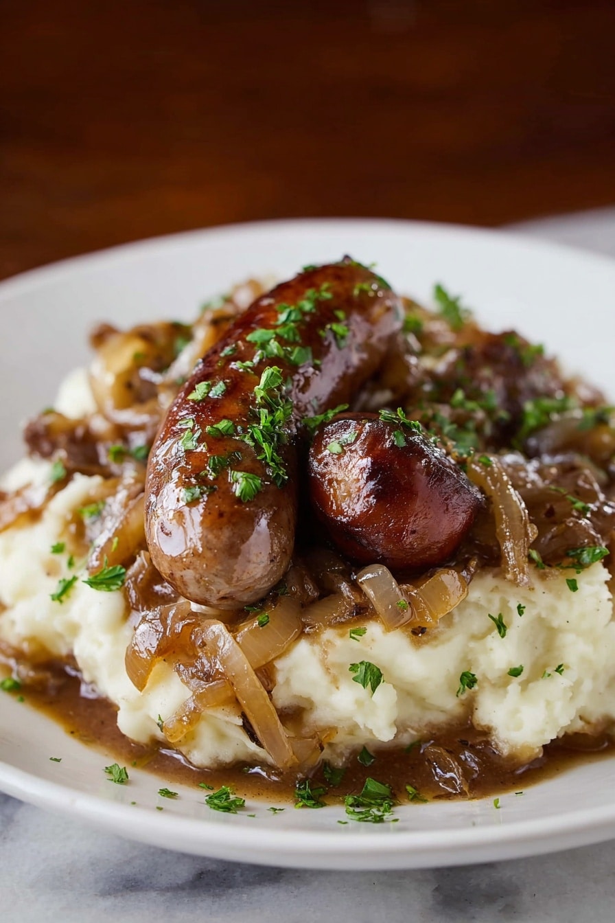 Stout Sausage and Mash with Onion Gravy Recipe 6 A white plate holds a mound of creamy mashed potatoes as the bottom layer, topped with rich brown gravy mixed with caramelized onions that appear soft and slightly translucent. On top, there are two browned sausages, one larger and one smaller, with a shiny, slightly crispy texture on the outside. The dish is sprinkled with fresh green chopped herbs for a pop of color. The setting has a white marbled surface and a deep brown blurred background, making the food stand out clearly. photo taken with an iphone --ar 2:3 --v 7 - Stout Sausage and Mash with Onion Gravy, hearty British pub dinner, comfort food recipes, savory sausage and mash, onion gravy with stout beer