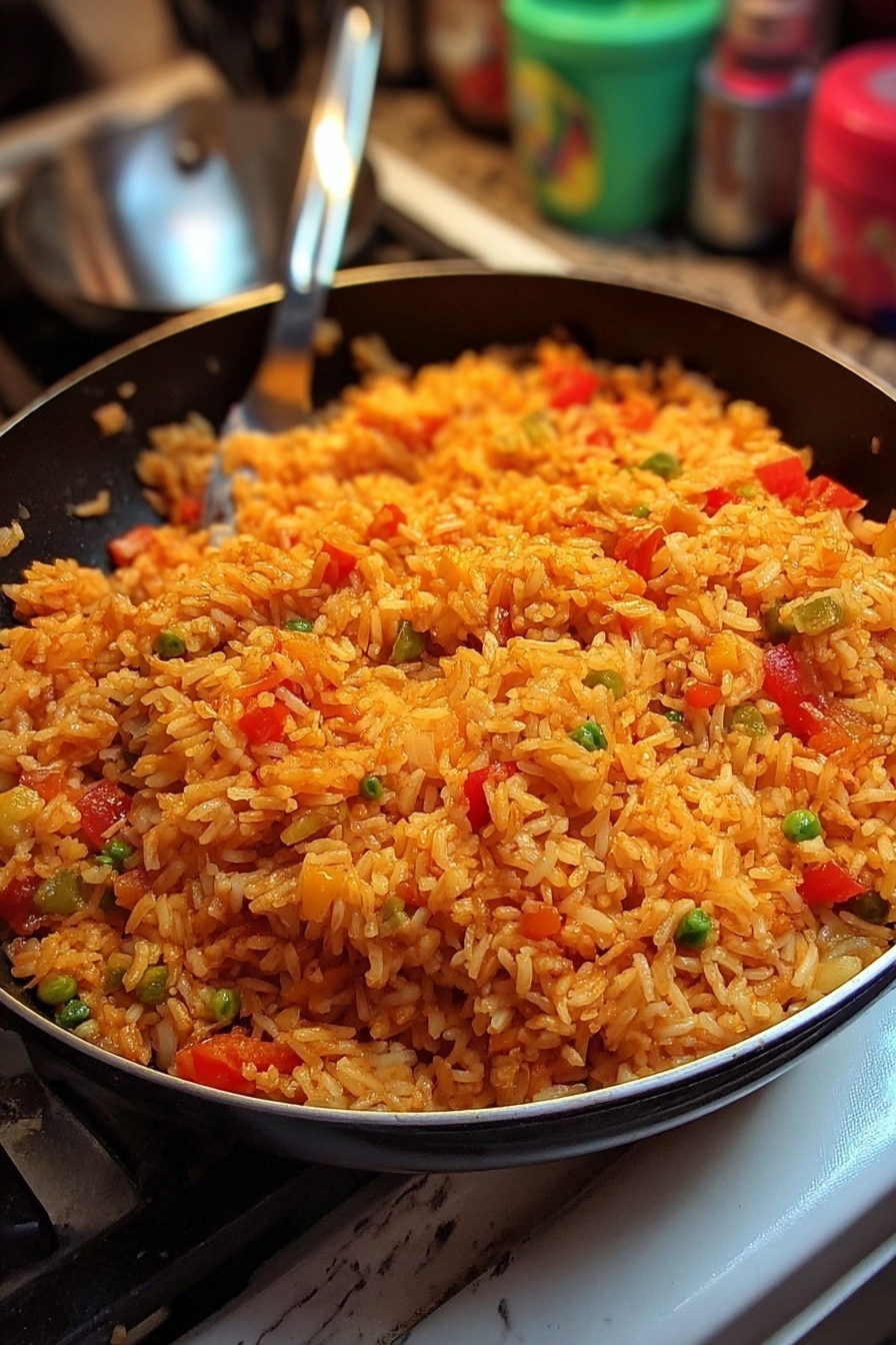 A large black pan filled with a mound of cooked rice that is orange in color, mixed with visible pieces of red bell pepper, green peas, and translucent cooked onions scattered throughout. The rice looks moist and fluffy with some spices giving it a rich texture. The pan is sitting on a stove with a white marbled surface in the background and some blurred colorful containers behind it. A silver cooking spoon is partially visible on the left side of the pan. Photo taken with an iphone --ar 2:3 --v 7 - Steak Queso Rice Bowl, how to make a steak queso rice bowl, easy steak rice bowl recipe, flavorful rice bowl with steak and queso, hearty steak quesadilla bowl