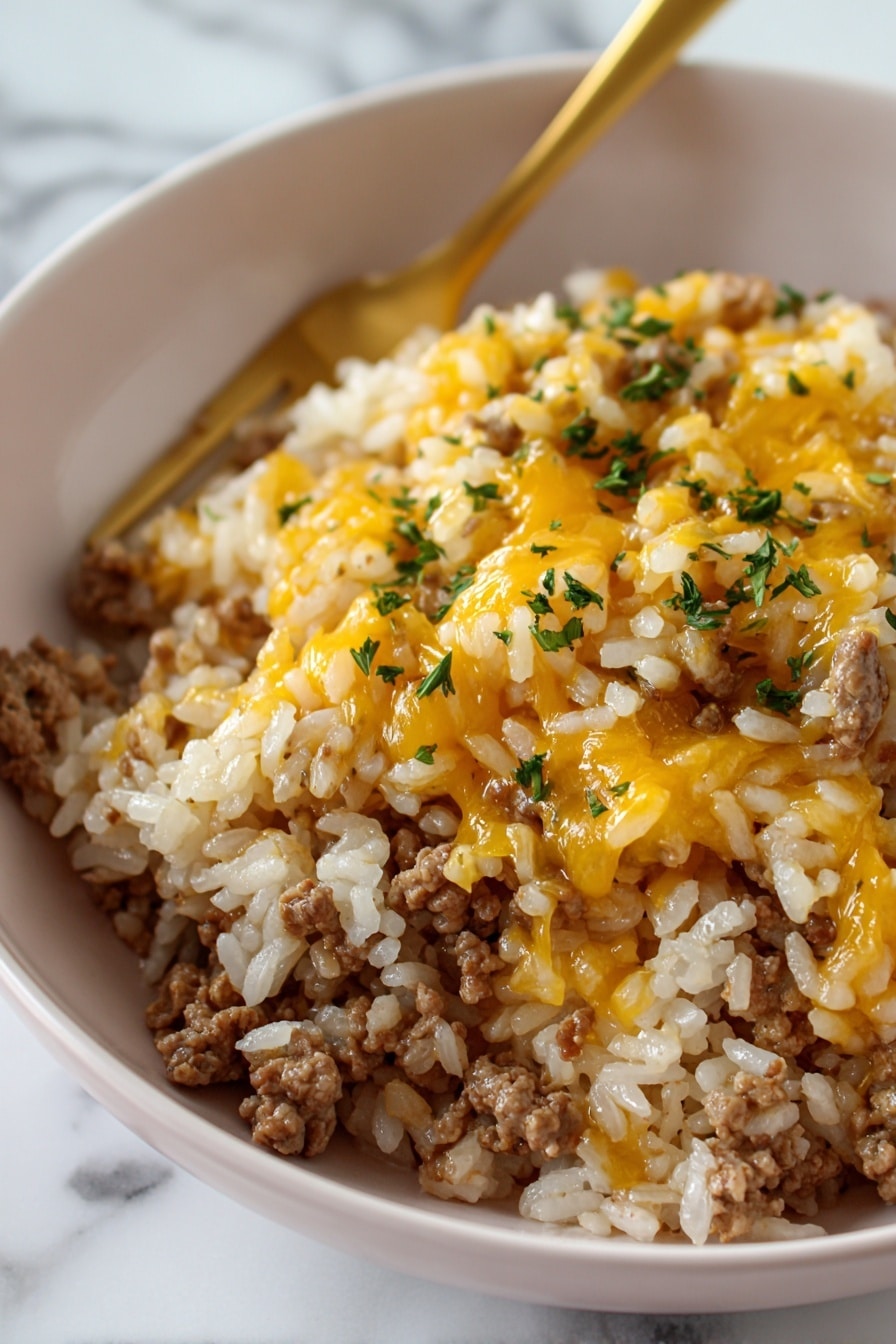 Cheesy Ground Beef and Rice Bake Recipe 8 The image shows a close-up of a white bowl filled with a single layer of cooked white rice mixed with browned ground meat and topped lightly with melted yellow cheese and small green parsley pieces scattered on top. In the background, a gold-colored fork is partially visible resting inside the bowl. The dish looks warm and textured, and the photo is taken on a white marbled surface. Photo taken with an iphone --ar 2:3 --v 7 - Cheesy Ground Beef and Rice Bake, easy ground beef casserole, cheesy beef and rice dish, weeknight dinner idea, comforting casserole recipe