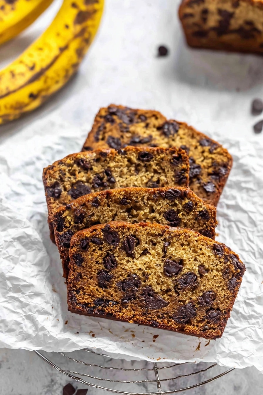 The image shows three slices of banana chocolate chip bread placed on crumpled parchment paper. The bread has a brown color with a moist texture and is filled with many dark chocolate chips spread evenly throughout. Behind the slices, part of a white wire cooling rack is visible. There is a yellow banana with brown spots in the top left corner on a white marbled surface. The overall scene is bright and clear, giving a fresh bakery feel. Photo taken with an iphone --ar 2:3 --v 7 - Moist Chocolate Chip Banana Bread, banana bread with chocolate chips, easy banana bread recipe, moist banana bread, best chocolate chip banana bread