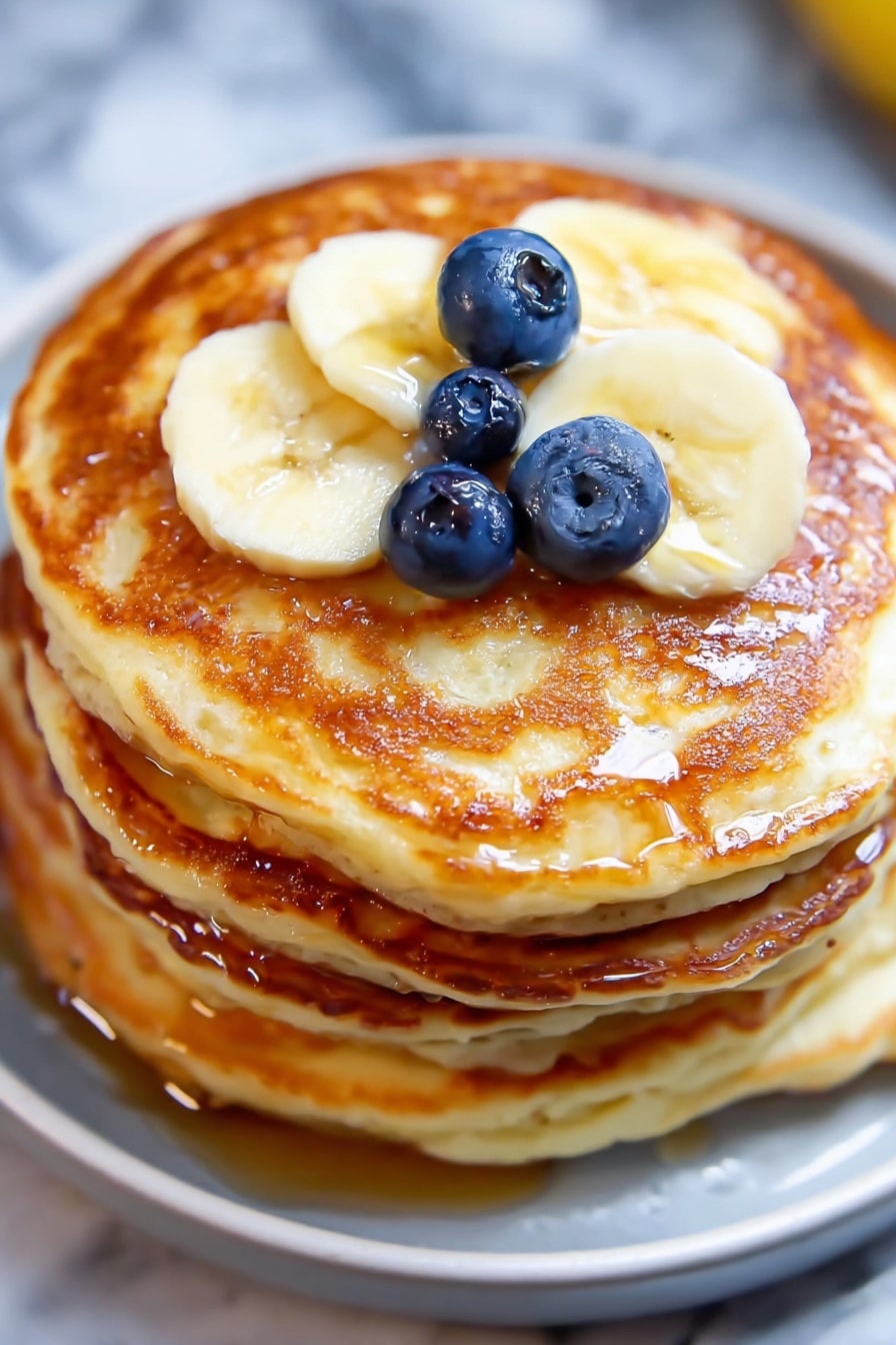 A stack of four thick, golden-brown pancakes with a slightly crispy texture on the edges is shown from a close view. On the top pancake, there are three round blueberries placed near the center, glistening slightly, and three slices of pale yellow banana arranged in a row towards the edge. A light drizzle of syrup adds a shiny, wet look on the top pancake. The stack sits on a white plate, all set on a white marbled surface. photo taken with an iphone --ar 2:3 --v 7 - Greek Yogurt Pancakes with Lemon, lemon yogurt pancake recipe, fluffy Greek yogurt pancakes, lemon breakfast pancakes, healthy Greek yogurt pancake